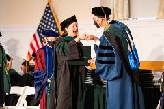 A woman receives her diploma and fist bump from the Dean. Both are wearing academic regalia.