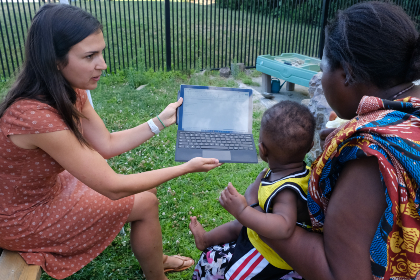 Mimi Falcone holds a laptop computer with screen facing a mother who has two young children on her lap.